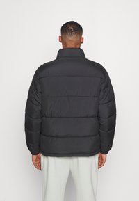 Giacca nera imbottita con colletto alto e cuciture orizzontali. Realizzata in materiale sintetico liscio con una texture leggermente riflettente.