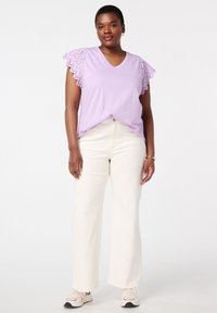 Haut violet à manches courtes avec un détail en dentelle festonnée sur les manches, assorti à un pantalon blanc à jambe large. Le modèle se tient sur un fond neutre.