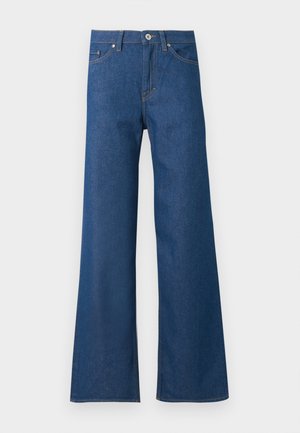 Sort kvinde iført en hvid skjorte med knapper over en hvid top, viddeblå jeans og sorte hælede sandaler, stående på en hvid baggrund.