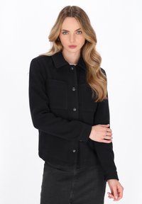 Jongeman met golvend blond haar, gekleed in een zwarte knopenjack en donkere jeans, poseert met één hand op haar arm tegen een witte achtergrond.