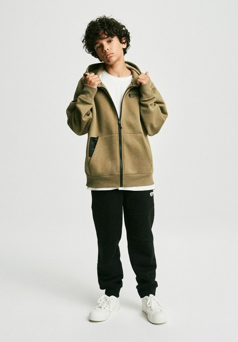 Kapturowa bluza polarowa w kolorze khaki z zapięciem na zamek i kieszeniami z przodu. Noszona z czarnymi spodniami dresowymi i białymi sneakersami. Gładki, casualowy design.