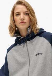Sudadera gris con cremallera y detalles en azul marino, con un cuello interior a rayas, cierre con cordón y logo bordado de "Superdry".