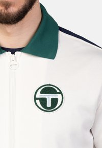 Weiße kapuzensportjacke mit marineblauen Akzenten, grünem strukturiertem Kragen und rundem, grünen und blauen gesticktem Logo auf der Brust.
