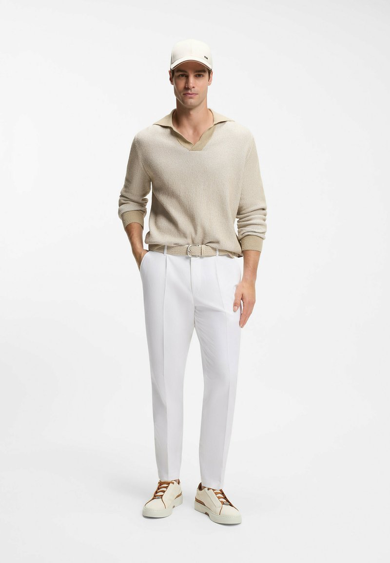 Homme portant un pull beige, un pantalon blanc, une ceinture assortie, une casquette beige et des baskets crème avec des lacets marron, debout devant un fond blanc.