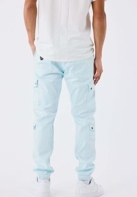 Uomo che indossa pantaloni cargo azzurro chiaro con polsini elasticizzati, sneakers bianche e una camicia bianca, in piedi contro uno sfondo semplice.