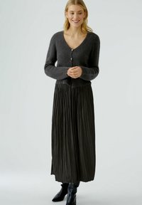 Houten cardigan met V-hals, knoopsluiting en lange mouwen, gecombineerd met een geplooide zwarte maxi-rok en zwarte enkellaarsjes.
