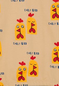 Tessuto beige con illustrazioni ripetitive di galline gialle con creste rosse e testo blu che dice "EARLY BIRD". La trama è liscia e morbida.
