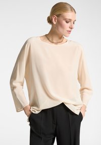 Blusa beige chiaro a maniche lunghe realizzata in un tessuto morbido e fluido, con scollatura tonda e vestibilità rilassata, abbinata a pantaloni neri.