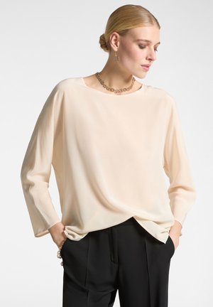 Blusa beige chiaro a maniche lunghe realizzata in un tessuto morbido e fluido, con scollatura tonda e vestibilità rilassata, abbinata a pantaloni neri.