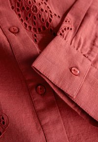 Punane linane pluus, millel on tikandiga kaunistatud avad, nööbitav krae ja mansetid, sile tekstuur ning peened värvivarjundid.