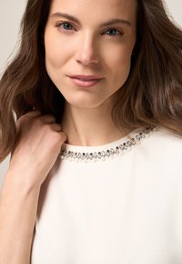 Prenda beige con escote redondo adornada con embellishments de perlas y cristales, con una textura suave y una silueta ajustada.