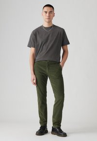 Joven de pie frente a un fondo sencillo, vistiendo una camiseta gris oscuro, pantalones verdes oliva, zapatos negros y un collar de cadena de plata.