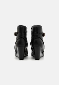 Botas de tobillo de cuero negro con un tacón alto y grueso, que presentan correas cruzadas y un cierre de cremallera en la parte posterior. Textura suave y diseño elegante.