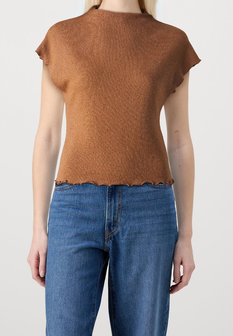 Kvinde iført en brun kortærmet strikbluse med en let bølget kant og blå denimjeans, stående mod en ensfarvet lys baggrund.
