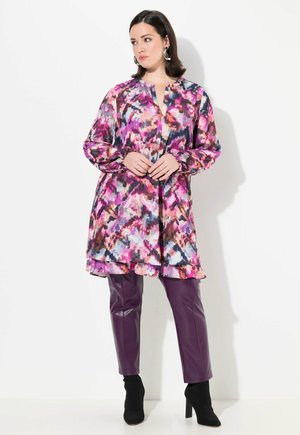 Farbige, mit abstrakten Mustern gestaltete Tunika-Bluse in Rosa und Lila, kombiniert mit figurbetonten lila Lederhosen und schwarzen Ankle Boots.