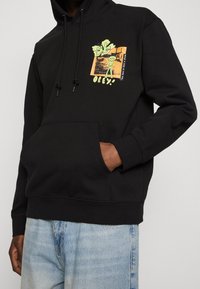 Haut à capuche noir avec une poche avant, un cordon de serrage pour la capuche et un design graphique coloré avec une figure verte et du texte sur la poitrine.