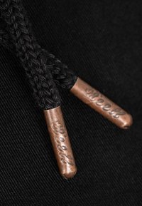 Primer plano de los extremos de un cordón de calzado trenzado negro rematados con terminales de cobre grabados con la palabra "Street" sobre un fondo de tela negra.