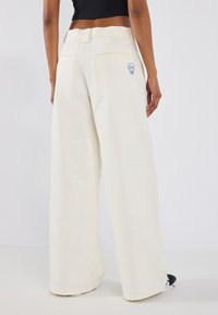 Pantalons larges en tissu crème, avec poches arrières et un graphique souriant bleu sur le côté droit. Texture lisse et coupe décontractée.