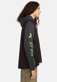 Schwarze ärmellose Weste über einem dunklen Kapuzenpullover, mit grünen botanischen Grafiken auf dem Ärmel. Lässige Passform mit weicher Textur.