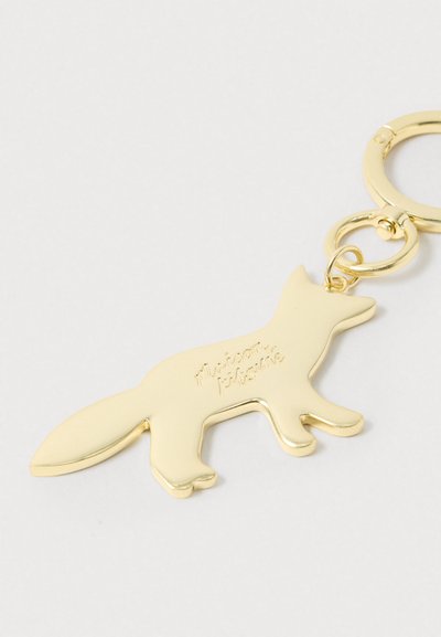 Μπρελόκ από χρυσό μέταλλο σε σχήμα αλεπούς με την επιγραφή «maison kitsuné» στο σώμα του, σε απλό λευκό φόντο.