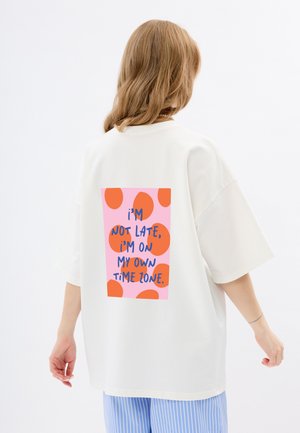 Witte oversized T-shirt met een kleurrijke print op de achterkant, met roze en oranje stippen en blauwe tekst die een speelse boodschap weergeeft.