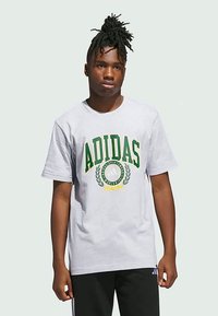 Jaunas vyras su pinto plauku vilki balta Adidas marškinėliais su žaliu ir geltonu logotipu, bei juodomis Adidas treniruočių kelnėmis prieš monotonišką foną.