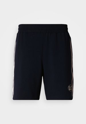 Tmavě modré sportovní šortky z měkkého materiálu, s elastickým pasem, bílými bočními pruhy a kulatým logem ve spodní pravé části.