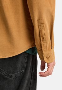 Braunes Cordhemd mit geknöpften Manschetten und strukturiertem Oberflächen, drüber getragen über einem grünen Hemd, kombiniert mit einer schwarzen Hose.