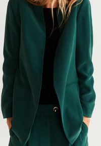 Blazer su misura color teal in tessuto liscio, con chiusura a un bottone e due tasche frontali. Presenta una vestibilità rilassata e un design minimalista.