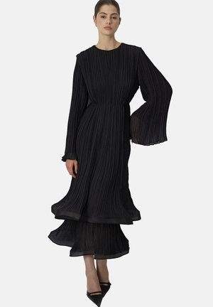Kvinde iført en lang sort plisseret kjole med udslåede ærmer og lagdelt kant, kombineret med sorte spidse hæle, stående mod en hvid baggrund.