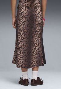 Person stående iført en brun og sort leopardmønstret nederdel, hvide sokker og mørkebrune snørestøvler foran en ensfarvet grå baggrund.