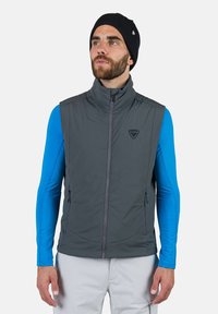 Graue gepolsterte Weste mit hohem Kragen, Vorderreißverschluss und seitlichen Taschen. Getragen über einem blauen Langarmshirt. Mit Logo auf der Brust.