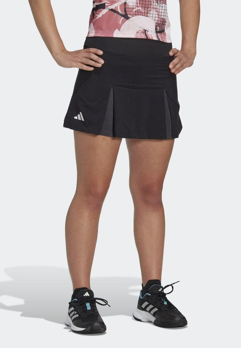 Jupe de sport noire à plis, en tissu léger. Présente un logo sur la hanche gauche. Portée avec des baskets noires et blanches.