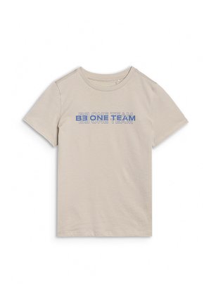 Enfant blond à la peau claire et aux taches de rousseur porte un t-shirt beige avec le texte « BE ONE TEAM » et un short bleu, debout devant un fond uni.