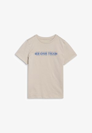 Enfant blond à la peau claire et aux taches de rousseur porte un t-shirt beige avec le texte « BE ONE TEAM » et un short bleu, debout devant un fond uni.