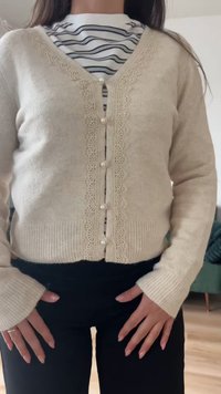 Γυναίκα με μπεζ πλεκτή ζακέτα με δαντελένιο τελείωμα και κουμπιά από πέρλες, πάνω από μαύρο-άσπρο ριγέ πουκάμισο, συνδυασμένη με μαύρο παντελόνι.