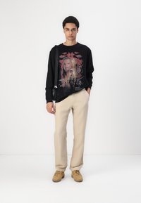 Cămașă neagră cu mâneci lungi, prezentând un imprimeu grafic colorat, asortată cu pantaloni bej deschis și adidași bej. Include o geantă simplă pentru umăr.