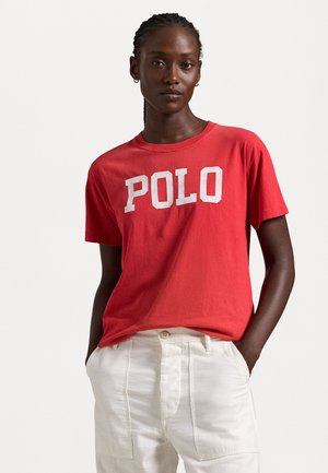 Red cotton T-shirt featuring a large white "POLO" print, round neckline, and short sleeves, paired with light-colored trousers.