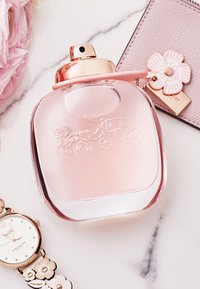 Clear glass perfume bottle with a pink tint, rounded shape, and gold cap. Features an embossed horse carriage design and sits beside a floral watch.