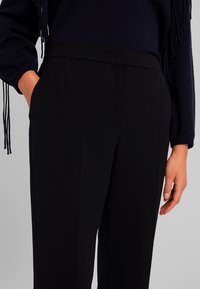 Pantaloni neri su misura con una texture liscia, caratterizzati da una parte anteriore piatta, tasche laterali e un design aderente. Accentuati da una leggera linea di piega.