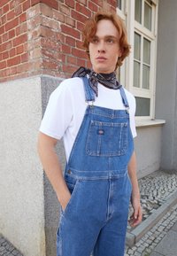Világoskék farmer overall, állítható vállpántokkal, első zsebbel és oldalzsebekkel. Fehér pólóval és fekete sállal viselve.
