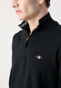 Crni pulover s patentnim zatvaračem, rebrastim ovratnikom, malim izvezenim logom na lijevoj strani prsa i teksturiranim pletenim uzorkom.