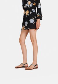 Abito floreale nero con maniche lunghe, vita stretta e orlo corto; abbinato a sandali piatti a thong con un accento metallico.