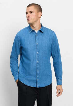 Uomo che indossa una camicia di jeans blu a maniche lunghe con bottoni e pantaloni neri, guarda verso sinistra con le mani rilassate.