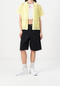 Žena oblečená v žlutém krátkém rukávovém háčkovaném kardiganu, bílém zkráceném tílku, černých šortkách ke kolenům, bílých ponožkách a bílých vysokých teniskách.