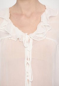 Blouse blanche semi-transparente avec col froufrou et fermetures à nouer à l’avant portée par une personne visible du cou à la poitrine.