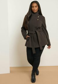 Bruine broekjas met hoge kraag, dubbele knoopsluiting en lange mouwen. Model draagt zwarte broek en enkellaarsjes, staand tegen een neutrale achtergrond.