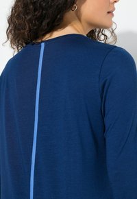 Laivastonsininen pitkähihoinen paita kaarevalla pääntiellä ja pystysuoralla vaaleansinisellä korostuksella takana. Pehmeä kangas, jossa hieman tekstuuria.