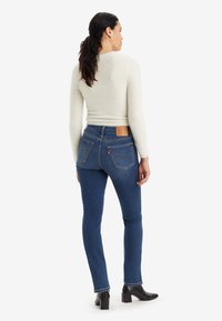 Jeans skinny in denim blu con tasche posteriori e un'etichetta in cuoio marrone sulla vita, abbinati a un top lungo a maniche lunghe di un leggero color crema strutturato.