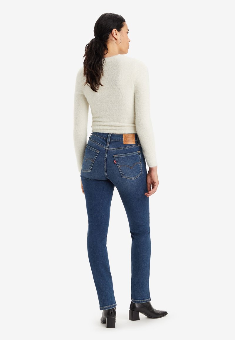 Jean skinny en denim bleu avec poches arrière et une étiquette en cuir marron à la taille, accompagnés d'un haut à manches longues texturé de couleur crème claire.